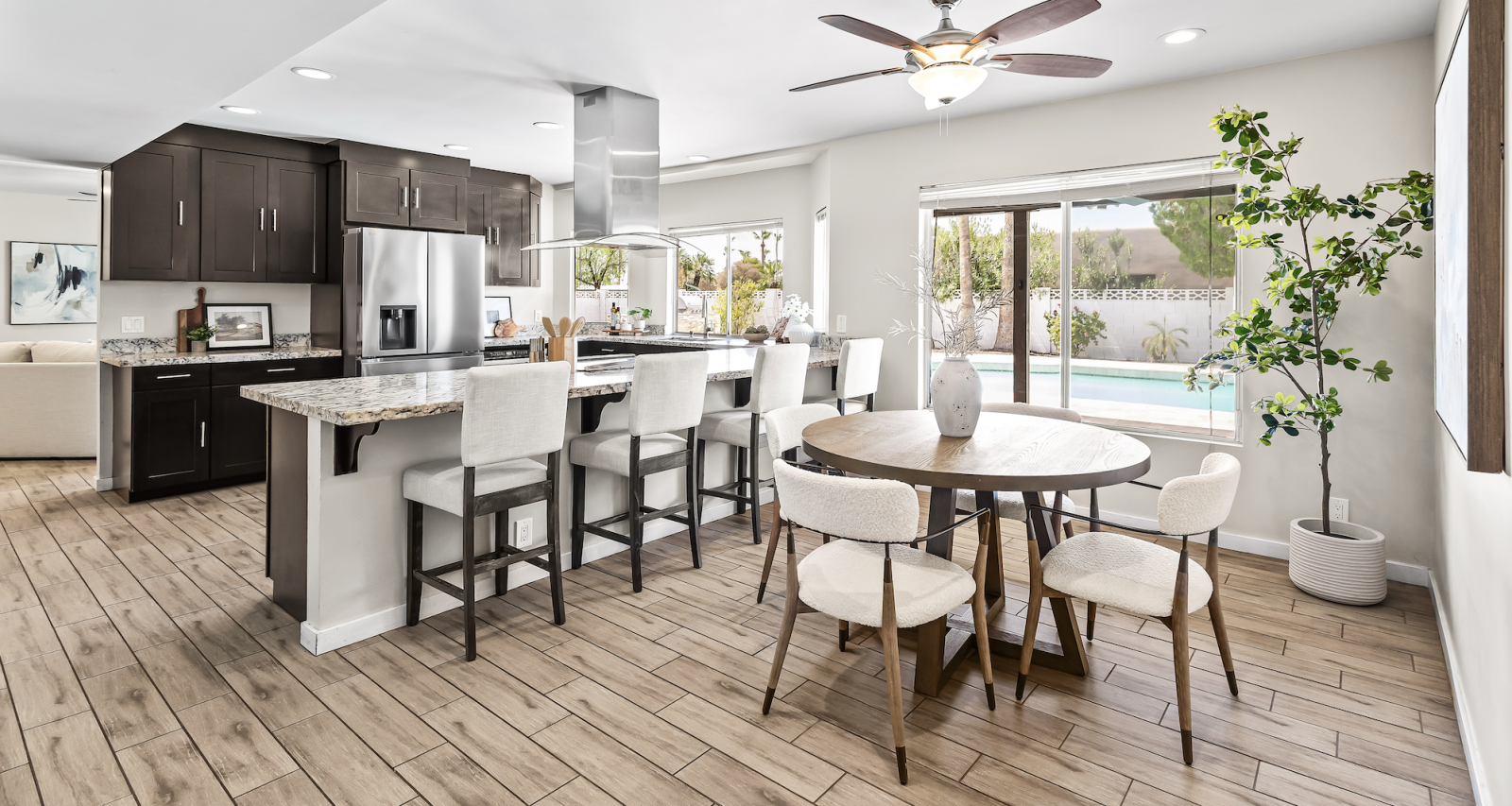 Open-concept kitchen with dark wood cabinets, granite countertops, and a marble-topped island, adjacent to a dining table by a window overlooking a backyard pool.